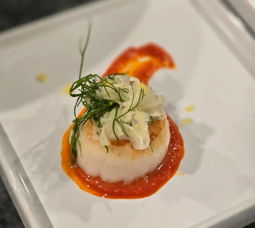 A seared scallop topped with shaved fennel and herbs on a pool of red sauce.
