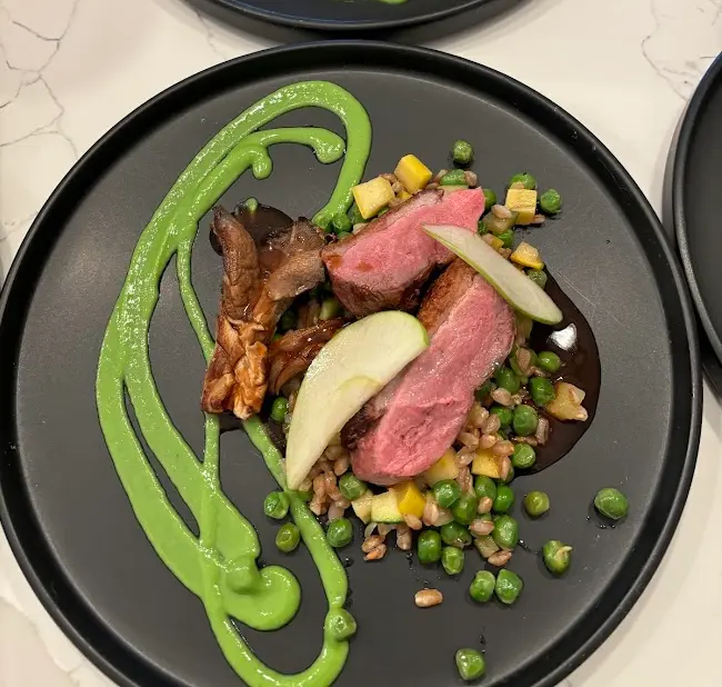 Sliced duck breast with spring peas, mushrooms and green puree on a black plate.