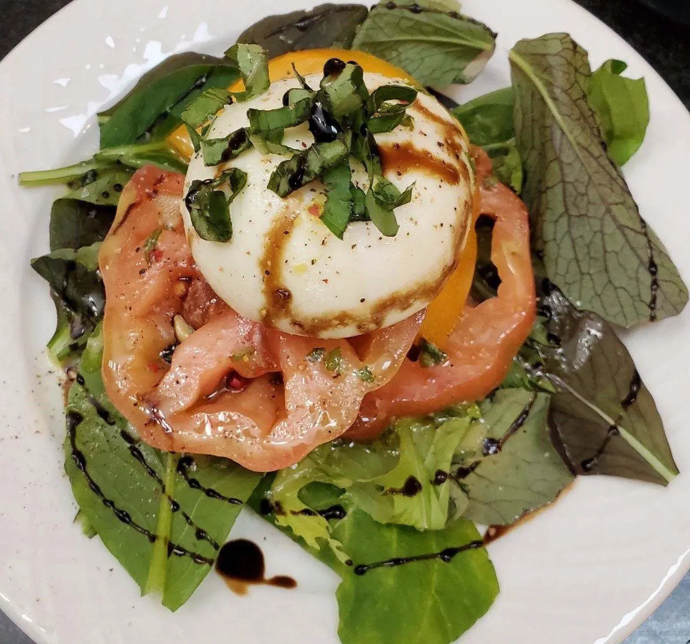 Vegan burrata-style cheese over tomato slices and leafy greens with a dark drizzle.