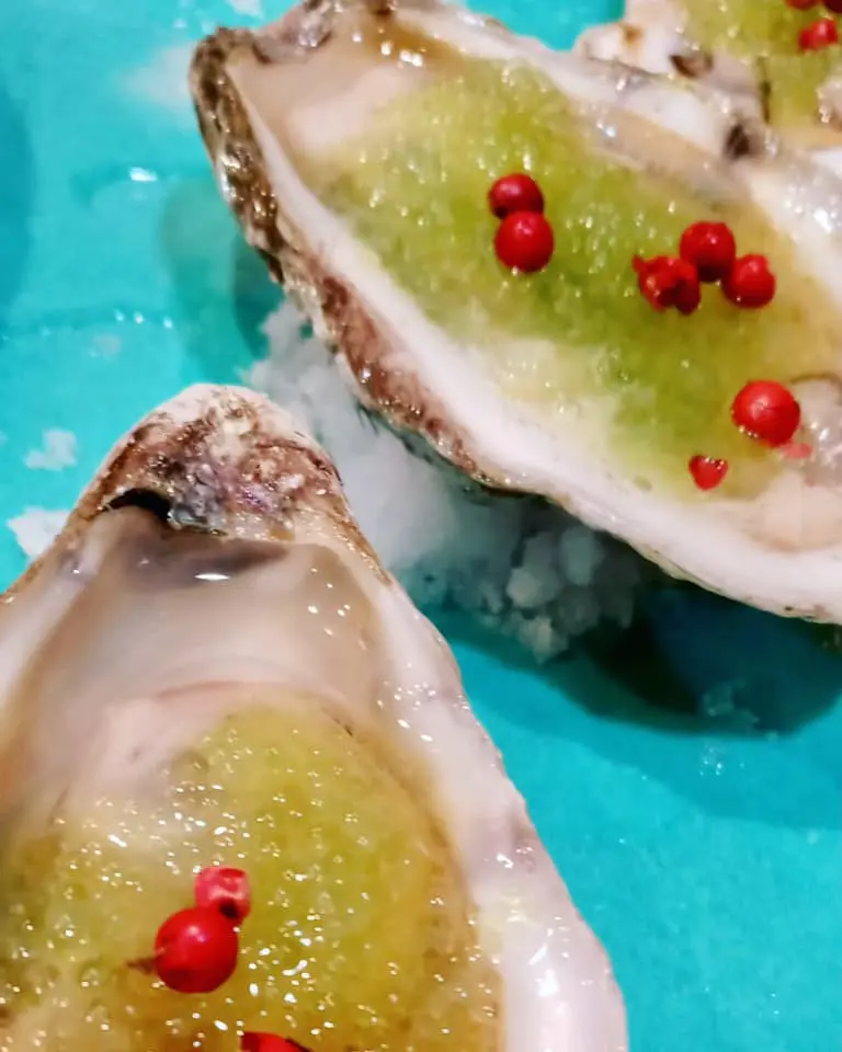 Freshly shucked oysters with green granita and small red garnishes on a blue platter.