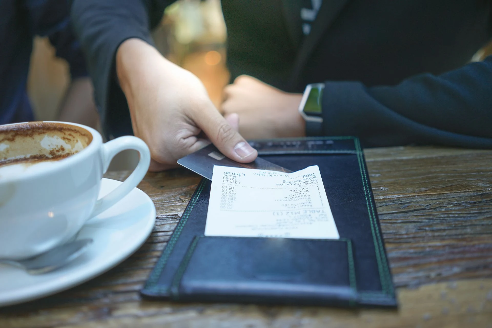 A restaurant bill in a check presenter beside a coffee cup and seated guest.