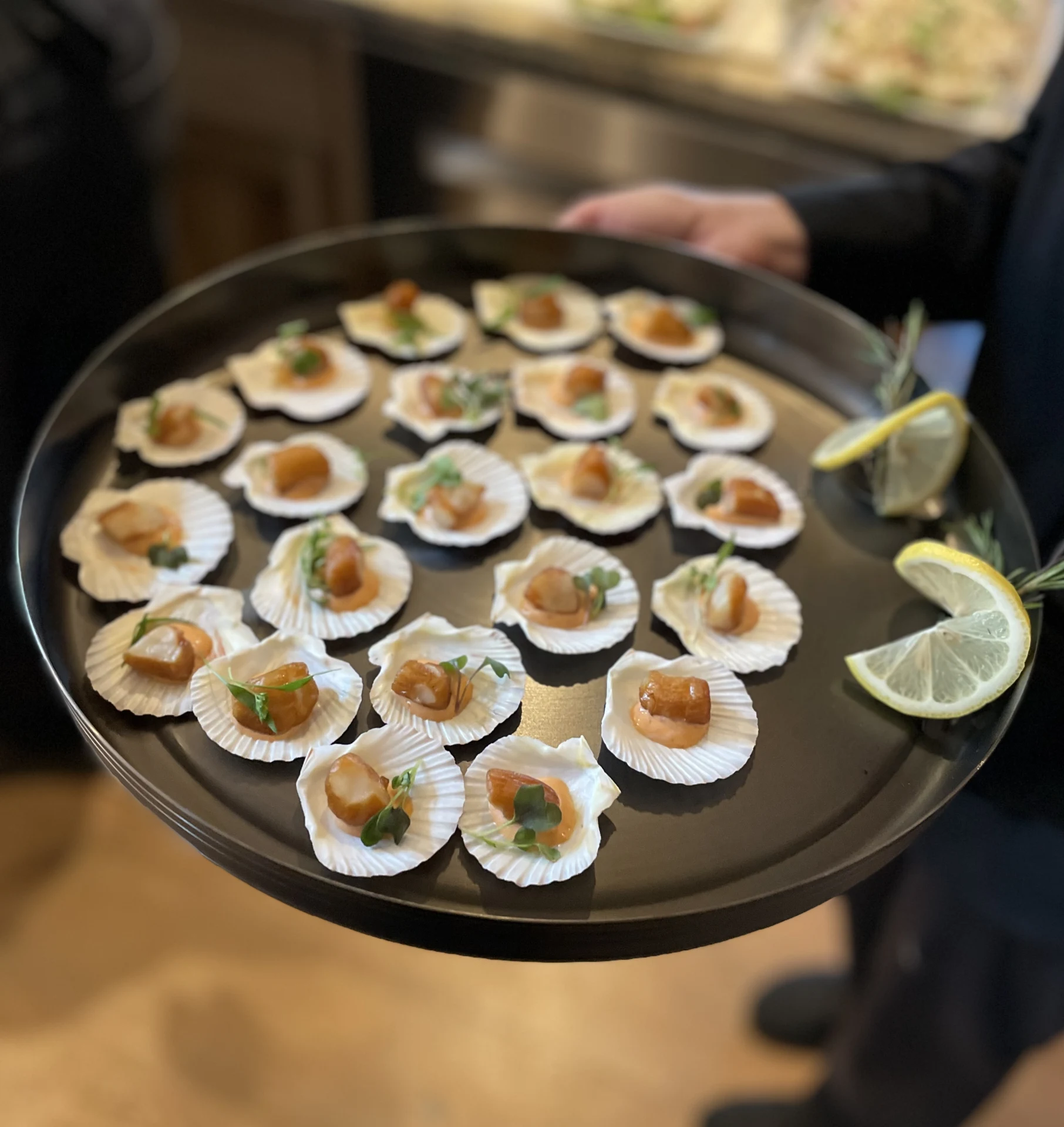 A steward carrying smoked bay scallop appetizers in shells on a round tray.