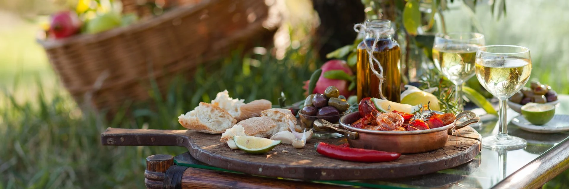 Fresh produce, cheeses and wine on an outdoor table beside a vegetable garden.